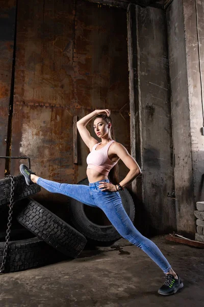 A dark-haired woman coach in a sporty  pink  short top and gym leggings  stretch   in an old garage, against the background of tires. Conceptual Sports Photo