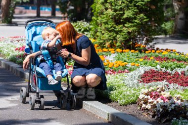 Genç bir anne bir bebek arabasıyla yürür. Bir yaşındaki oğlu sıcak bir yaz gününde şehir parkında neşeyle gülümser ve öpüşür.