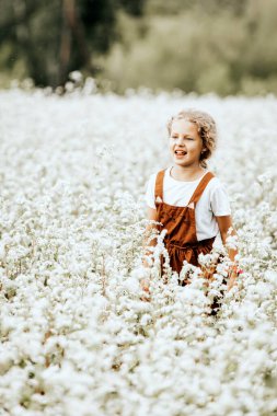 Kahverengi elbiseli güzel bir kızın portresi beyaz çiçeklerle tarlada koşuyor, güneşli bir yaz gününde günün tadını çıkarıyor. Çocuğun ve doğanın birliği kavramı, huzurlu ruh hali, çevre hayatı.