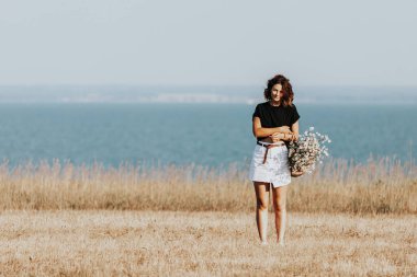 Sarı tarlada yürüyen, siyah t-shirt ve etek giyen, elinde bir buket papatya ile hasır bir çanta tutan genç bir kadın.