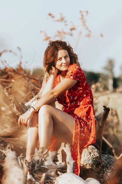 Açık hava atmosferik yaşam tarzı fotoğrafı. Kırmızı elbiseli güzel, siyah saçlı bir kadın. Sahanın tepesinde, arka planda, günbatımlı bir ormanda oturuyor..
