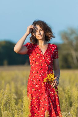 Kırmızı romantik elbiseli, sarı çiçekli güzel bir kadının portresi. Güneşli bir yaz gününde, günün tadını çıkarıyor. Çocuğun ve doğanın birliği kavramı, huzurlu ruh hali, çevre hayatı.