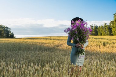 Güzel elbiseli güzel bir kadın, siyah şapkalı, mor çiçekli büyük bir buket, gün batımında bir çayırda gülüyor ve dışarıda yürüyor. Livestyle yürüyüş konsepti