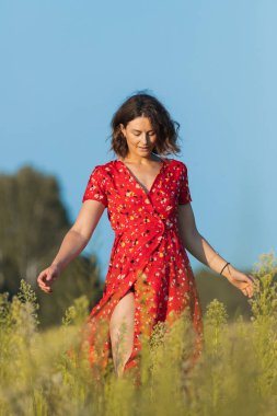 Tarlalar boyunca kırmızı uzun elbiseli kıvırcık saçlı bir kızın romantik yürüyüşü. Kadın özgürlüğü, özgürlük ve aşk kavramı.