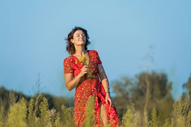 Yeşil tarlada yürüyen ve elinde bir buket sarı çiçekle kırmızı elbisesi olan genç bir kadın. Köyde yaz tatili ve yaşam tarzı kavramı.
