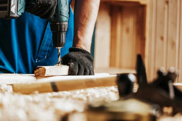 Close-up of a male construction worker in a black dirty construction overall closes a screw with a black modern screwdriver in a wooden block in the workshop