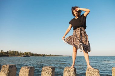 Kıvırcık saçlı, elbisesiyle denizde taş üstünde poz veren bir kadının romantik yürüyüşü. Kadın özgürlüğü, özgürlük ve aşk kavramı.