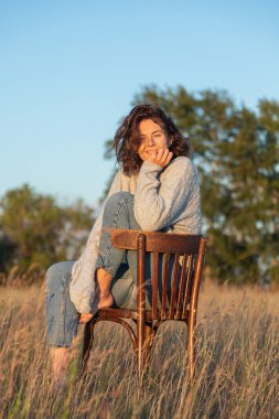 Kıvırcık saçlı, kot pantolonlu ve örgü ören bir kadının eski bir retro sandalyede arka planda oturduğu romantik bir yürüyüş. Kadın özgürlüğü, özgürlük ve aşk kavramı.