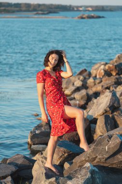 Kıvırcık saçlı, kırmızı elbiseli, denizde taş üstünde poz veren bir kadının romantik yürüyüşü. Kadın özgürlüğü, özgürlük ve aşk kavramı.