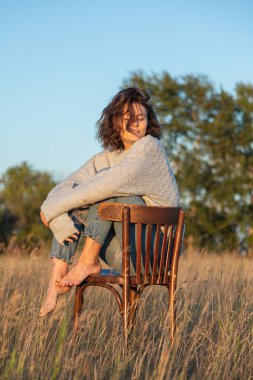 Kıvırcık saçlı, gülümseyen ve yan bakan romantik bir kadının ön görüntüsü. Eski bir sandalyede oturup güneşli bir yaz gününde çayırda dinleniyor.