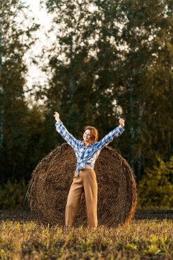 Ekose gömlekli, kıvırcık saçlı, samanlıkta gülümseyen ve dans eden ve güneşli bir yaz gününde çayırda dinlenen romantik bir kadın.