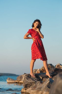 Kıvırcık saçlı, kırmızı elbiseli bir kadının, gökyüzü ve denizin arka planına karşı bir taşın üzerinde romantik yürüyüşü. Kadın özgürlüğü, özgürlük ve aşk kavramı.
