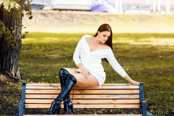 İnce vücutlu, güzel, siyah saçlı bir kadın beyaz elbiseli, yüksek topuklu bir bankta Freen City Park 'a karşı oturuyor.