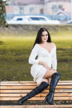 Beyaz, siyah saçlı, romantik elbiseli genç bir kadın bir yaz günü şehir parkında bir bankta oturuyor.