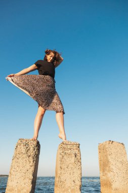 Siyah tişörtlü ve etekli kıvırcık saçlı bir kadının denizde taş üstünde durduğu romantik bir yürüyüş. Kadın özgürlüğü, özgürlük ve aşk kavramı.