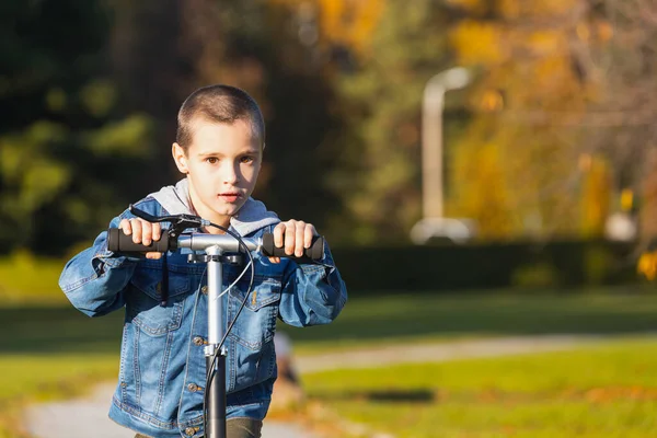 デニムジャケットの陽気な男の子はすぐに都市公園のスクーターにロールします 暖かい秋の日に歩く — ストック写真
