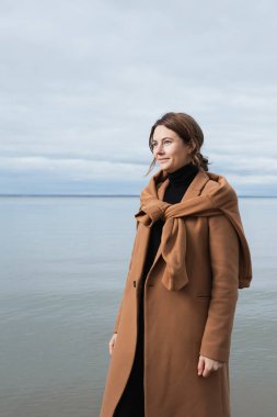 Güneşli bir sonbahar gününde denize karşı poz veren bej renkli yün ceketli güzel bir mankenin portresi. Sonbahar sıcacık fotoğrafı. Kadın gülümsüyor ve kameraya bakıyor, neşeli bir ruh hali..