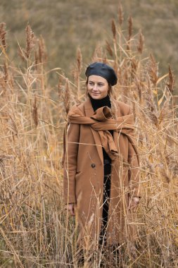 Siyah bereli güzel genç bir mankenin portresi ve bej renkli yün bir palto güneşli bir sonbahar gününde arka planda günün tadını çıkarır. Kadınların ve doğanın birliği kavramı, barışçıl ruh hali.