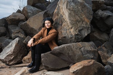 Sıcak giysiler içinde güzel bir genç modelin portresi güneşli bir sonbahar gününde arka planda kayalar ve deniz üzerinde günün tadını çıkarır. Sonbahar sıcacık fotoğrafı. Gülümseyen ve yüzünü çeviren, neşeli bir kadın..