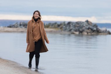 Sahilde yürüyen ve denize karşı sıcak kıyafetler giyen genç bir kadın. Köyde yaz tatili ve yaşam tarzı kavramı, Seascape
