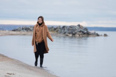 Bir sonbahar günü kumlu bir plajda mavi bir gökyüzü yakınında şık giysiler içinde genç bir kadının atmosferik portresi. Kadın özgürlüğü ve özgürleşme kavramı.