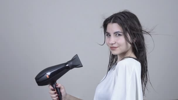 Mulher bonita segurando um secador de cabelo em um fundo cinza. Cuidados com o cabelo. — Vídeo de Stock