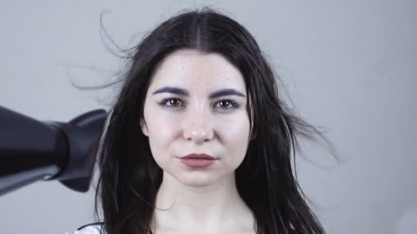 Όμορφη νεαρή γυναίκα που κρατά ένα στεγνωτήρα μαλλιών σε γκρι φόντο — Αρχείο Βίντεο