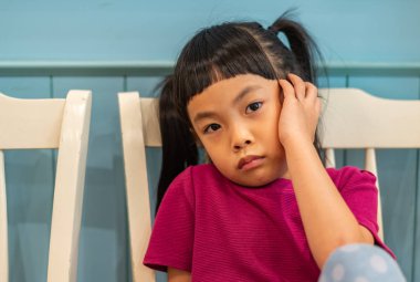 Moody little Asian girl, sitting and looking at camera. Wooden wall as backdrop. clipart