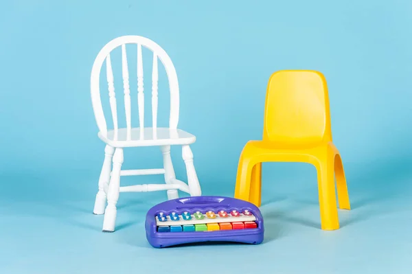 Small white wooden chair and yellow plastic chair for little children. Music instrument toy on floor. Soft color background.