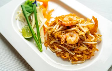 Pad Thai veya Tayland geleneği kızarmış erişteyi karıştırır. Beyaz tabakta karidesli Tayland eriştesi, yanında sebze ve limon, masada da doğal ışık..