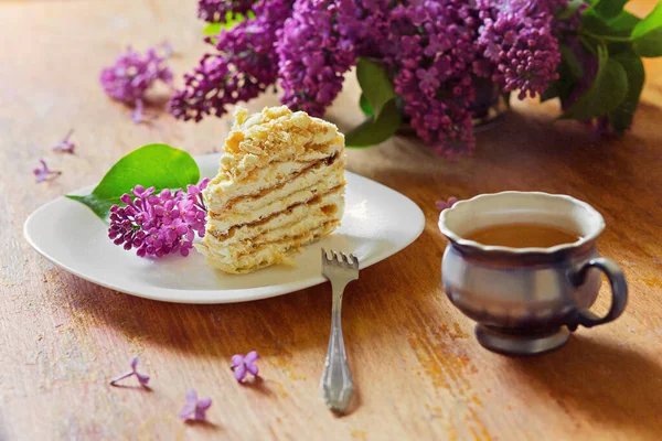A piece of cake lies on a plate with lilac flowers on the table and a mug of tea. Breakfast