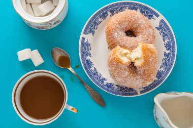 Bir fincan kahve, şeker ile bir tabak çörek ve mavi arka plan üzerinde diğer kahvaltı nesneleri kaplı.