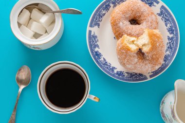 Bir fincan kahve, şeker ile bir tabak çörek ve mavi arka plan üzerinde diğer kahvaltı nesneleri kaplı.