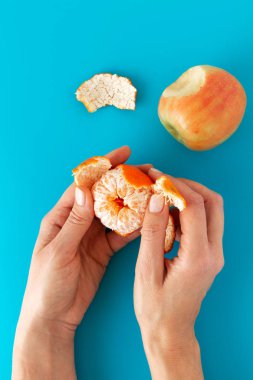 Bir mandalina peeling eller ve bir ısırık, mavi arka plan ile bir elma.
