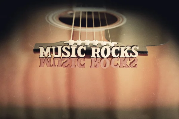 Cuvinte Muzică Rocks cu litere din lemn, pe suprafața reflectând o chitară acustică. Perspectiva podului chitarelor. Creative grungy retro fundal . — Fotografie, imagine de stoc