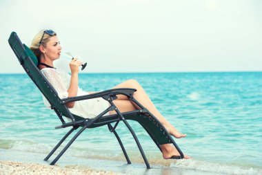 Genç kadın bir şezlong ve içme şarap kumsalda oturur.