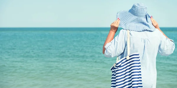An elderly woman in a hat looks at the sea. Summer vacation.