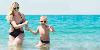 Güzel genç anne ve küçük oğlu sahilde dalgalar üzerinde çalışan elele. Eğlenceli, Aile, dost yaz tatili.