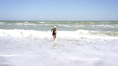 Siyah mayo ve hasır şapkalı genç seksi kadın sahilde güneşleniyor ve dinleniyor. Kız dalgalarda koşuyor..