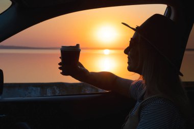 Genç bir kadın kahve içiyor ve gün batımında arabanın camından dışarı bakıyor..