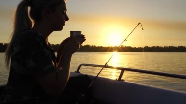 Bir kadın balık tutarken termostan çay içiyor. Bir tekneden gölde balık tutmak. Güneş ışığında olta kamışı. Balıkçılık konsepti.
