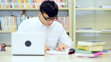 Kütüphane genç öğrenci çalışmada zor. Asyalı erkek üniversite öğrenci kitaplığı kitap ve Resepsiyon ve gülümseyen laptop ile çalışma araştırma yapıyor. Okul eğitim teknoloji kavramı için geri.