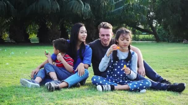 家庭吹泡泡在公园里 混合种族家庭与白人男子 中国妇女和他们的孩子 一起坐在公园里的草地上一起玩 — 图库视频影像