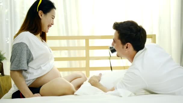 家族と親子関係の概念 妊娠中のアジア女性と笑って お互いに話して 遊んで 女の腹をなでる自宅ベッドの上に横たわる彼女の白の夫 — ストック動画