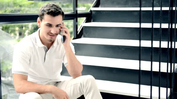 年轻英俊的白衣男子穿着白色衬衣和裤子坐在楼梯上 用手机和客户在现代办公室里聊天 穿着时髦的休闲服 靠在玻璃墙外的树木 — 图库视频影像