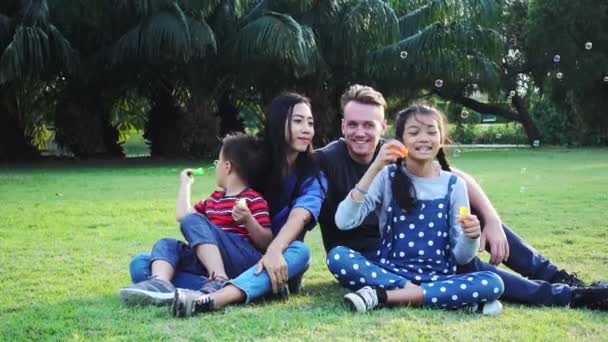 公園で家族の吹く泡 白人男性 中国人女性とその子供との混合人種家族 公園の芝生に一緒に座って楽しみながら — ストック動画