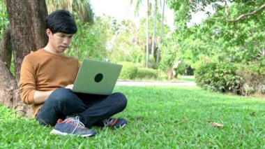 Parkta Laptop kullanan genç adam. Büyük bir ağacın yanında çim üzerinde oturan, rahatlatıcı ve bir dizüstü bilgisayar kullanarak. Kopya alanı ile.