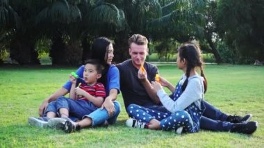 Aile parkta kabarcık üfleme. Beyaz adam, Çinli kadın ve çocukları ile karışık yarış ailesi. Park içinde çim birlikte eğlenmek birlikte oturmak.