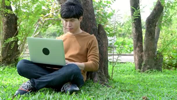 Молодой Человек Использует Ноутбук Парке Сидя Траве Рядом Большим Деревом — стоковое видео