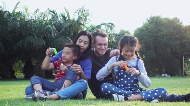家庭吹泡泡在公园里 混合种族家庭与白人男子 中国妇女和他们的孩子 一起坐在公园里的草地上一起玩 — 图库视频影像